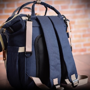 May include: A navy blue backpack featuring adjustable shoulder straps with beige webbing, a black mesh back panel, top carry handles, and a small detachable side pouch. The bag includes gold-toned zippers and metal hardware accents throughout.