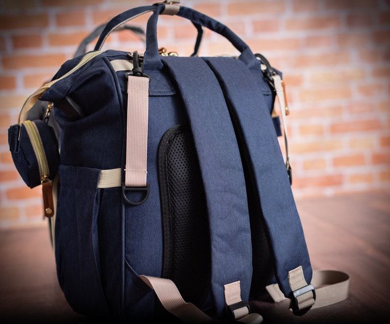 May include: A navy blue backpack featuring adjustable shoulder straps with beige webbing, a black mesh back panel, top carry handles, and a small detachable side pouch. The bag includes gold-toned zippers and metal hardware accents throughout.
