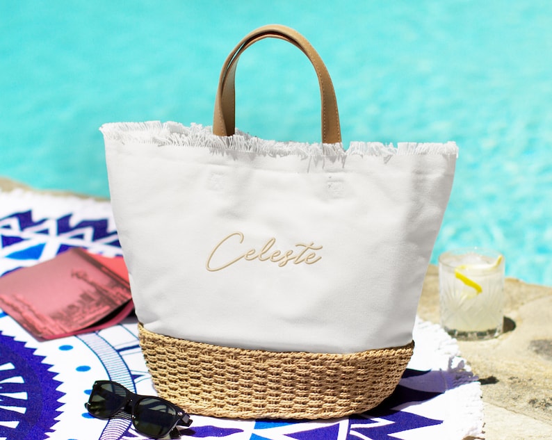 May include: White canvas tote bag with a brown leather handle and a woven straw bottom. The bag has a frayed edge and the name "Celeste" is embroidered on the front in a light brown thread.