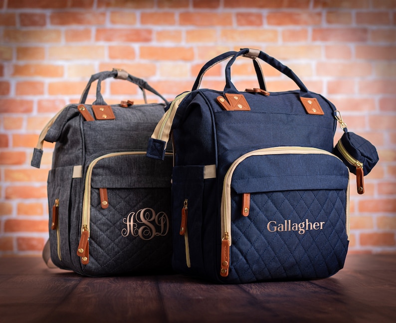 May include: Two quilted diaper bags with zippered pockets and monogrammed initials. The bag on the left is gray with the initials "HSD" and the bag on the right is navy blue with the name "Gallagher" embroidered on the front.