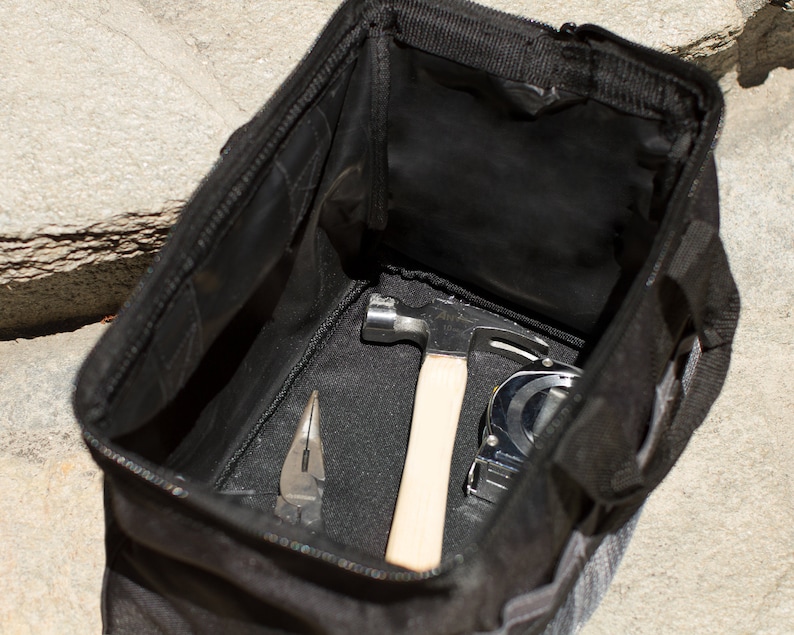 Puede incluir: Una bolsa de herramientas negra con una abertura con cremallera, que contiene un martillo, una cinta m&eacute;trica y un par de alicates.