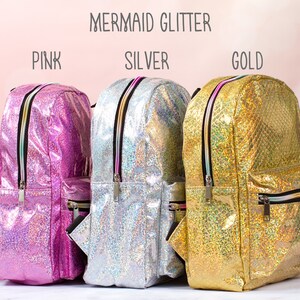 May include: Three iridescent glitter backpacks in pink, silver, and gold. The backpacks have a zippered front pocket and a top handle.