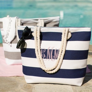 Puede incluir: Dos bolsas de lona a rayas con asas de cuerda. Una bolsa es gris y blanca, la otra es azul marino y blanca. La bolsa azul marino tiene un monograma bordado en la parte delantera. Ambas bolsas est&aacute;n sobre una toalla rosa.