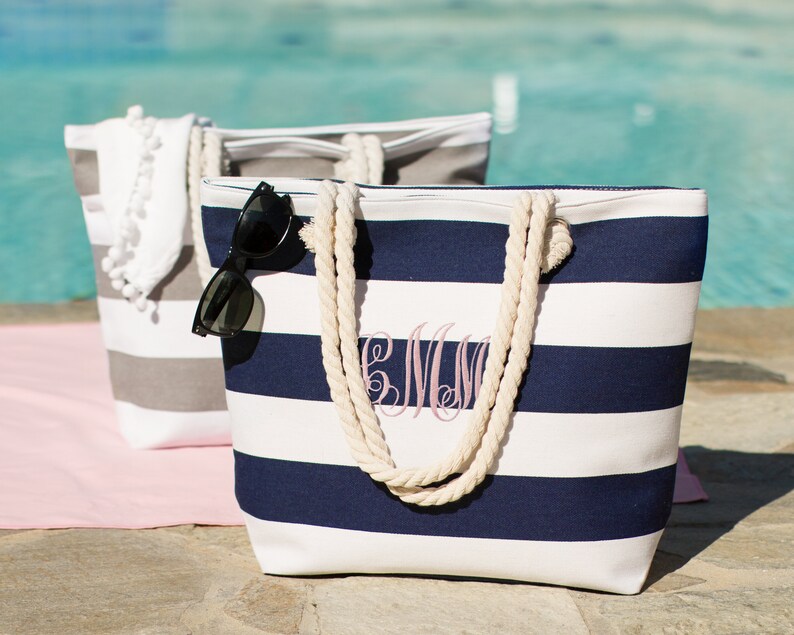 Puede incluir: Dos bolsas de lona a rayas con asas de cuerda. Una bolsa es gris y blanca, la otra es azul marino y blanca. La bolsa azul marino tiene un monograma bordado en la parte delantera. Ambas bolsas est&aacute;n sobre una toalla rosa.
