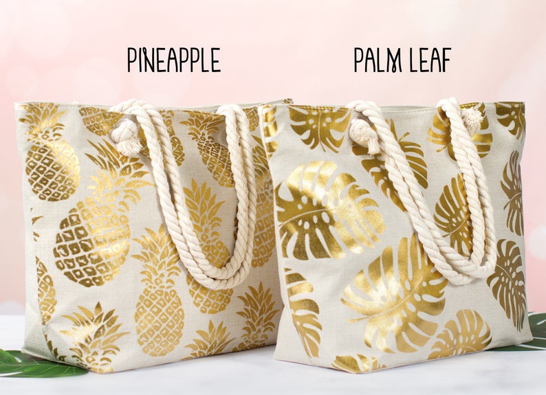 May include: Two canvas tote bags with rope handles. The bag on the left has a gold pineapple pattern on a beige background. The bag on the right has a gold palm leaf pattern on a beige background. The text "Pineapple" is above the left bag and the text "Palm Leaf" is above the right bag.