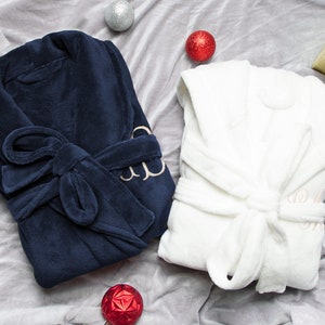 May include: Two plush bathrobes, one navy blue and one white, with embroidered initials. The robes are tied with matching sashes and are displayed on a gray background.