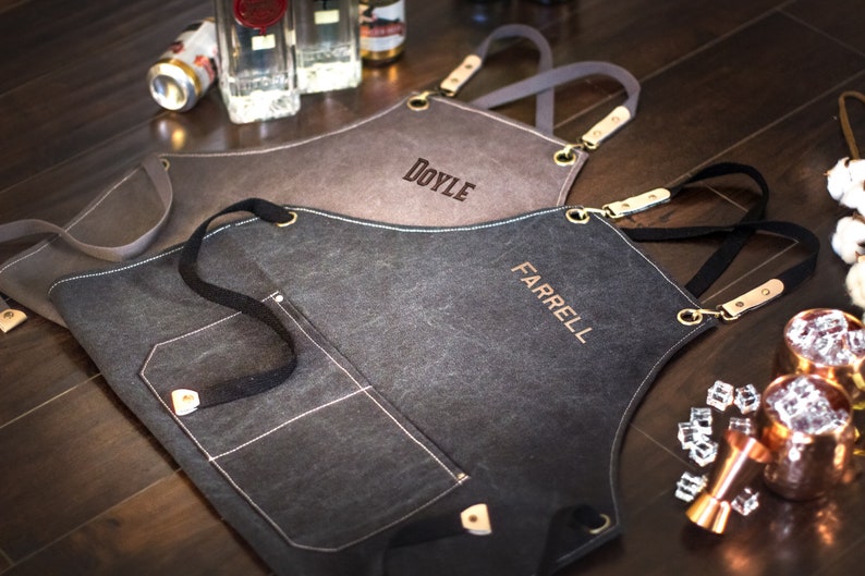 May include: Two personalized black canvas aprons with adjustable straps and a large pocket. The aprons are embroidered with the names "DOYLE" and "FARRELL".