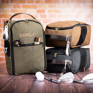 May include: A green canvas golf bag with the name "BARCLAY" embroidered on the front. The bag is sitting on a wooden surface with two brown and black golf bags stacked on top of each other. There are golf balls and golf clubs in the image.