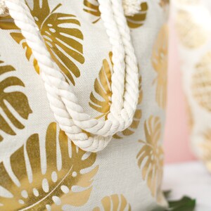 May include: A white canvas tote bag with gold foil monstera leaf print and white rope handles.