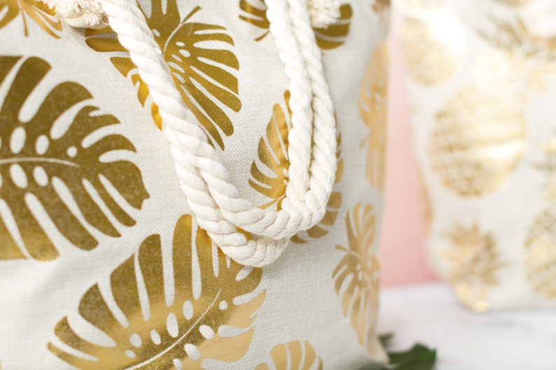 May include: A white canvas tote bag with gold foil monstera leaf print and white rope handles.