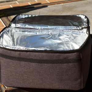 May include: A gray insulated lunch bag with a zippered closure and a black strap. The bag is open and the inside is lined with silver foil.