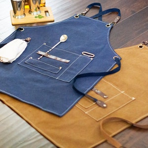May include: Two canvas aprons with adjustable straps, one blue and one brown. Both aprons have multiple pockets and are designed for use in a bar or kitchen.