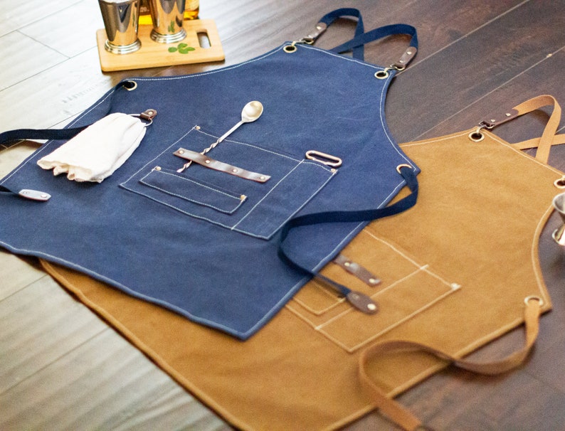 May include: Two canvas aprons with adjustable straps, one blue and one brown. Both aprons have multiple pockets and are designed for use in a bar or kitchen.
