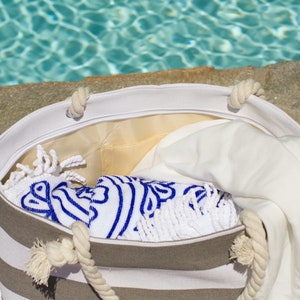 May include: A striped beach tote bag with rope handles, filled with a white towel and a blue and white patterned towel. The bag is open, revealing a yellow interior lining. The scene is set near a pool.