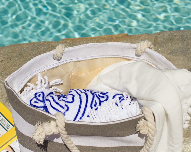 May include: A striped beach tote bag with rope handles, filled with a white towel and a blue and white patterned towel. The bag is open, revealing a yellow interior lining. The scene is set near a pool.