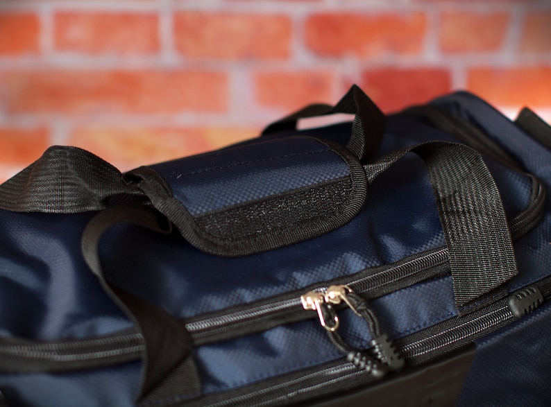 May include: A navy blue duffel bag with black straps and zips. The bag has a small pocket on the top with a black zip.