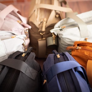 May include: An assortment of duffel bags in various colors, including light pink, gray, black, navy blue, tan, and orange. Each bag features a top handle, a shoulder strap, and gold-colored zippers.