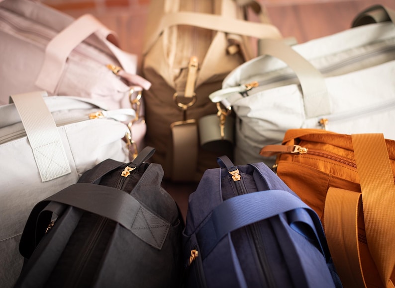 May include: An assortment of duffel bags in various colors, including light pink, gray, black, navy blue, tan, and orange. Each bag features a top handle, a shoulder strap, and gold-colored zippers.