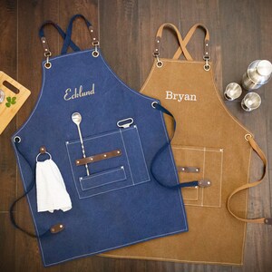 May include: Two personalized canvas aprons, one blue and one brown, with leather straps and pockets. The blue apron has a bar spoon and a bottle opener in the pocket. The brown apron has the name "Bryan" embroidered on it. The blue apron has the name "Ecklund" embroidered on it.