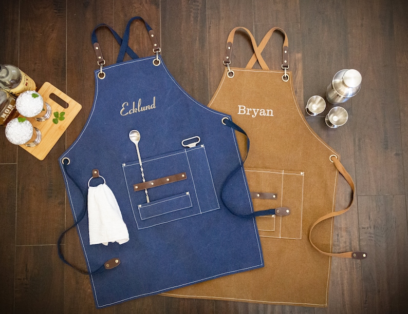May include: Two personalized canvas aprons, one blue and one brown, with leather straps and pockets. The blue apron has a bar spoon and a bottle opener in the pocket. The brown apron has the name "Bryan" embroidered on it. The blue apron has the name "Ecklund" embroidered on it.