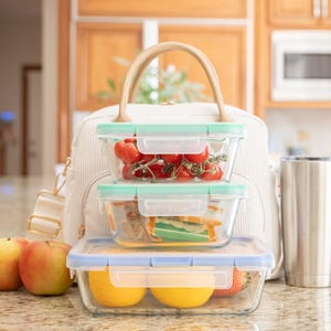 May include: A cream-colored lunch bag with a tan handle sits behind three glass food containers filled with fresh produce. Two apples and a stainless steel tumbler are in front of the bag. The containers hold tomatoes, sandwiches, and fruit.