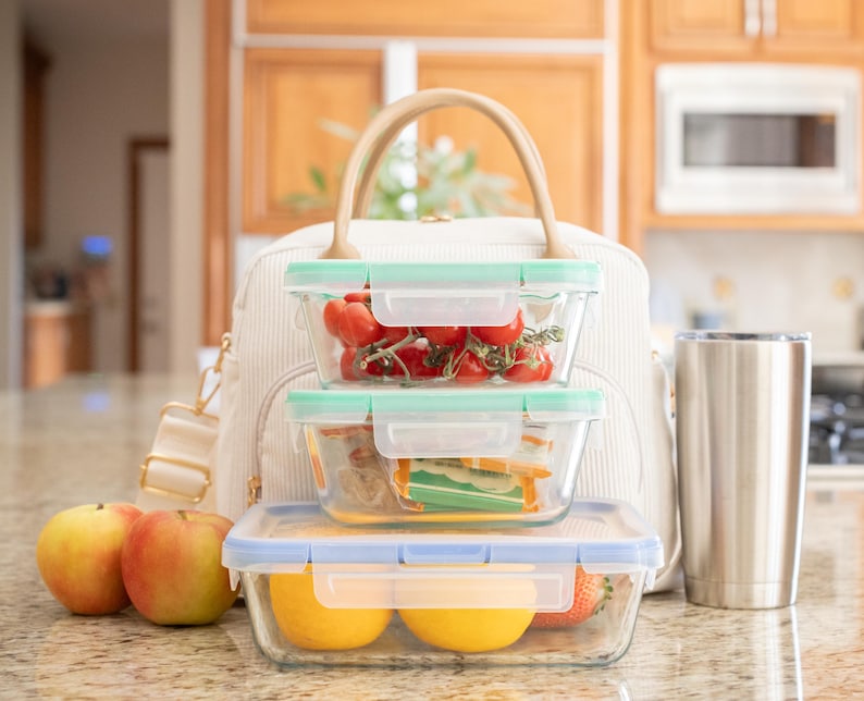 May include: A cream-colored lunch bag with a tan handle sits behind three glass food containers filled with fresh produce. Two apples and a stainless steel tumbler are in front of the bag. The containers hold tomatoes, sandwiches, and fruit.