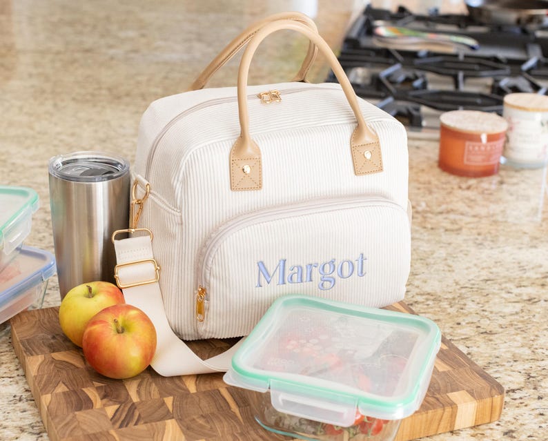 May include: Cream-colored corduroy lunch bag with a light brown handle and strap. The bag has a front pocket with the name "Margot" embroidered in blue. Includes a stainless steel tumbler, two apples, and a clear food container.