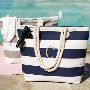 Puede incluir: Dos bolsas de lona a rayas con asas de cuerda. Una bolsa es gris y blanca con pompones blancos. La otra bolsa es blanca y azul marino con la letra "C" bordada en la parte delantera.