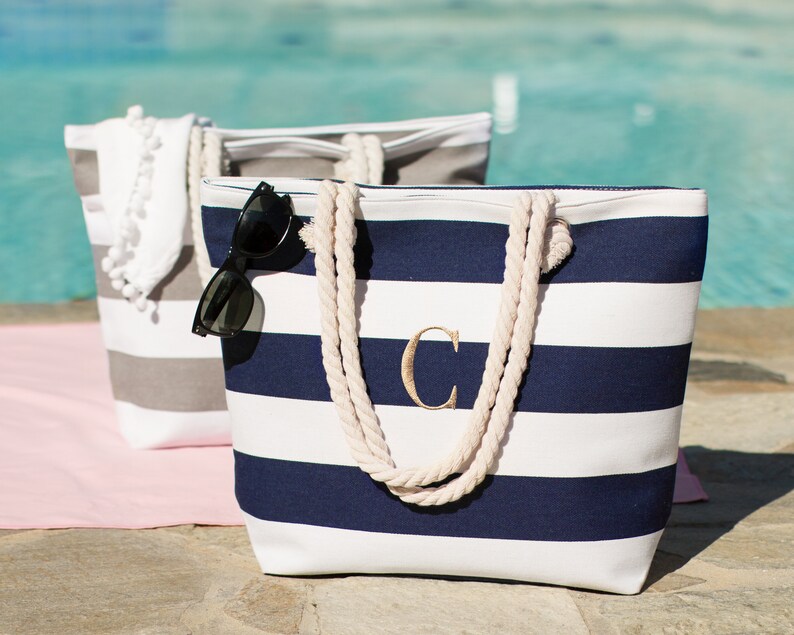 Puede incluir: Dos bolsas de lona a rayas con asas de cuerda. Una bolsa es gris y blanca con pompones blancos. La otra bolsa es blanca y azul marino con la letra "C" bordada en la parte delantera.
