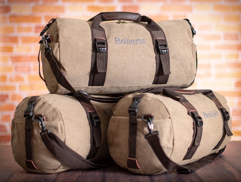 K&ouml;nnte beinhalten: Drei braune Canvas-Reisetaschen mit braunen Ledergriffen und -riemen. Die Taschen sind mit den Namen "Roberts", "Williams" und "Benfield" personalisiert.
