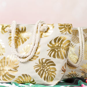 May include: Two beige canvas tote bags with gold leaf tropical leaf designs. The bags have rope handles.