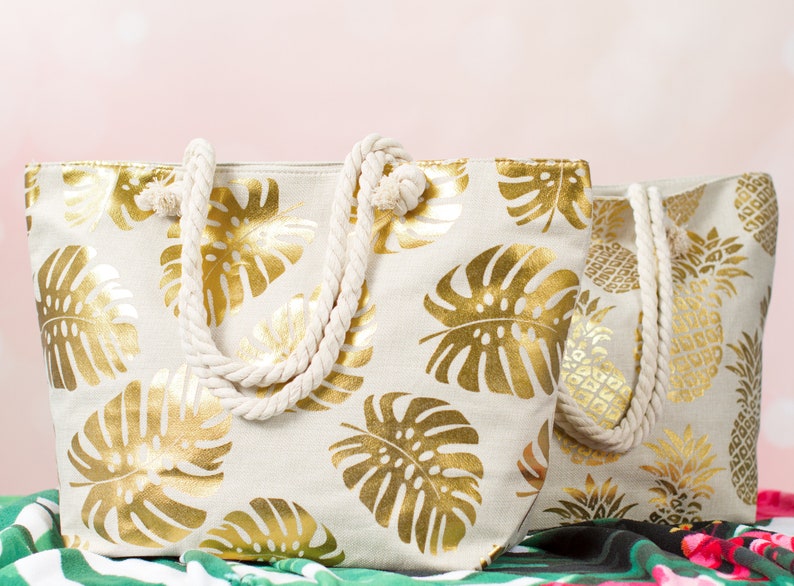 May include: Two beige canvas tote bags with gold leaf tropical leaf designs. The bags have rope handles.