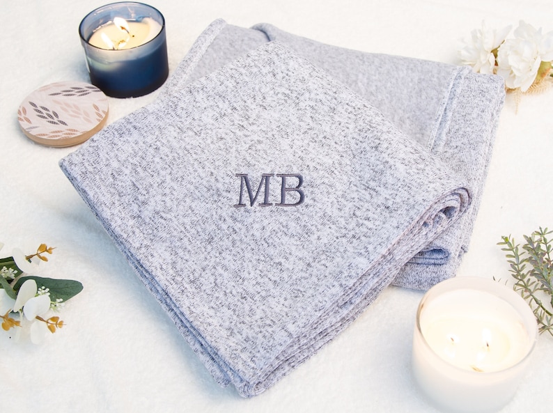 May include: A gray and white throw blanket with the initials "MB" embroidered in dark gray. The blanket is folded and displayed on a white surface with two candles.