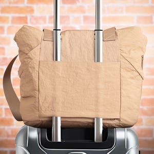 May include: A tan tote bag with a shoulder strap is secured to a silver rolling suitcase. The tote bag has a rectangular pocket on the front and is made of a soft, quilted material. The background is a brick wall.