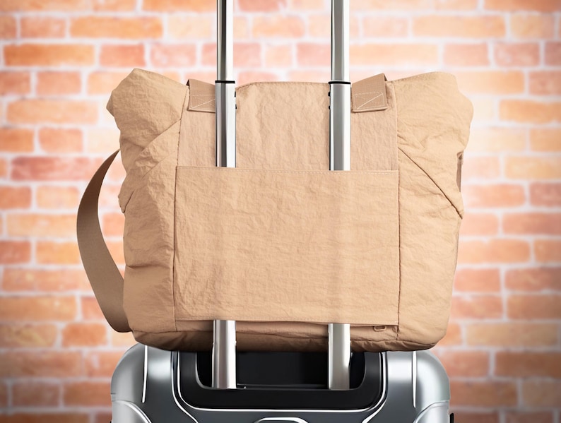 May include: A tan tote bag with a shoulder strap is secured to a silver rolling suitcase. The tote bag has a rectangular pocket on the front and is made of a soft, quilted material. The background is a brick wall.