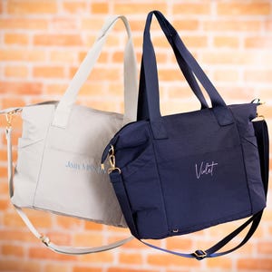 May include: Two tote bags, one light gray and one navy blue, with long handles and shoulder straps. The light gray bag has the text "Joan MSN, RN" embroidered in blue. The navy blue bag has the name "Violet" embroidered in white.