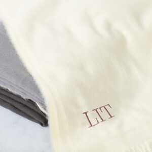 May include: A white throw blanket with a fringe border. The blanket is folded and has the initials "LIT" embroidered in a burgundy color.