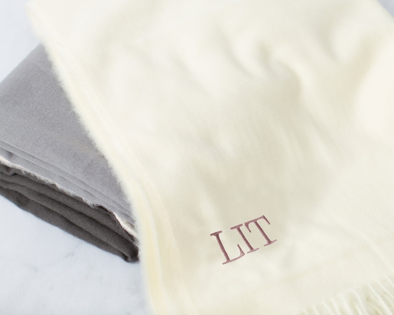 May include: A white throw blanket with a fringe border. The blanket is folded and has the initials "LIT" embroidered in a burgundy color.