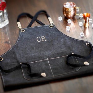 May include: Black canvas apron with adjustable straps and a large pocket. The apron has white stitching detail and the initials "CR" embroidered in gold thread.