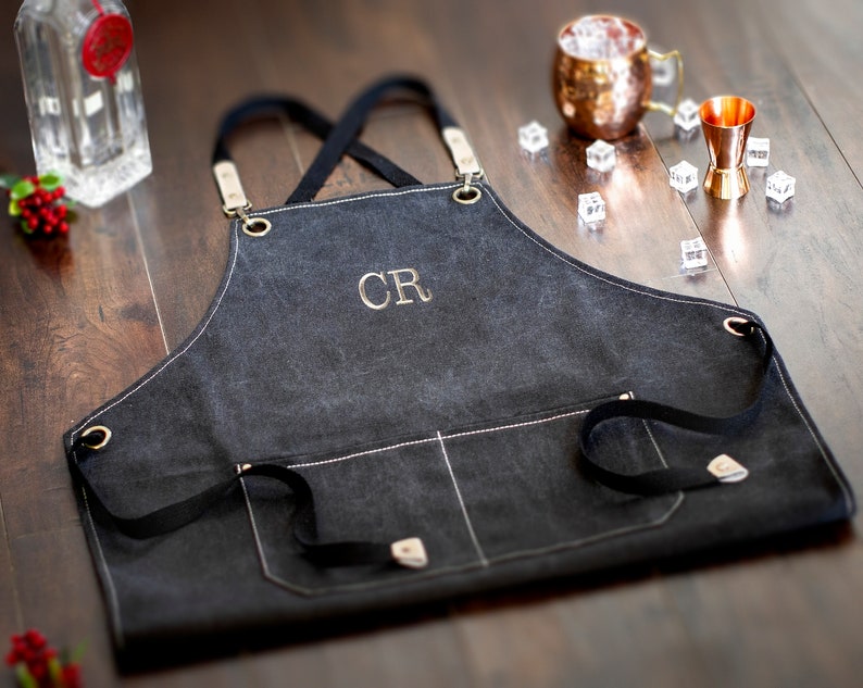 May include: Black canvas apron with adjustable straps and a large pocket. The apron has white stitching detail and the initials "CR" embroidered in gold thread.