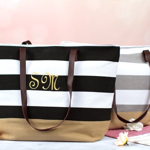 Puede incluir: Dos bolsas de lona a rayas con asas de cuero marr&oacute;n. La bolsa negra y blanca tiene el monograma "SM" bordado en la parte delantera. La bolsa gris y blanca es lisa.