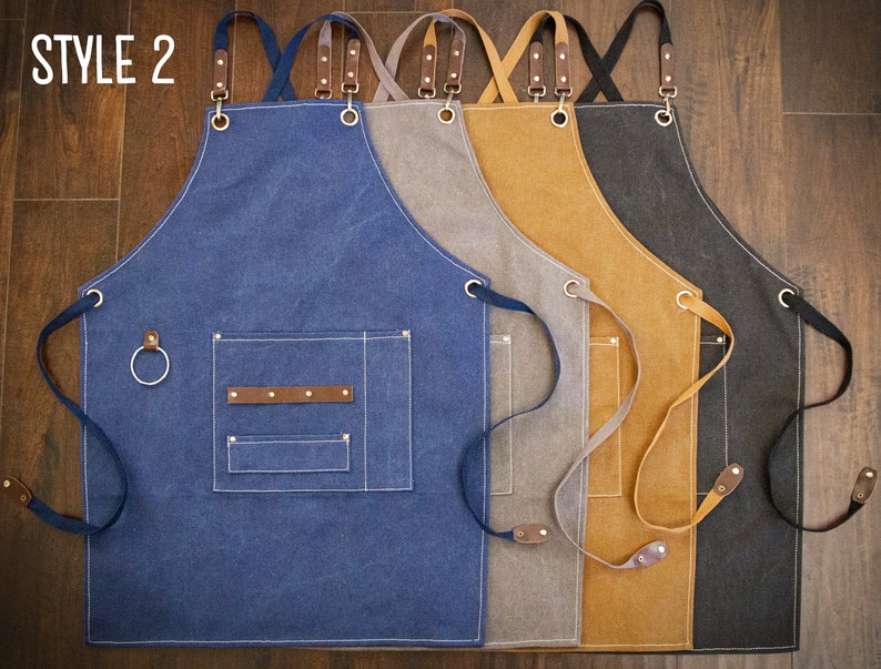 May include: Four canvas aprons in different colors: blue, gray, brown, and black. Each apron has adjustable straps, a large pocket, and a small loop on the side. The aprons are labeled "STYLE 2".