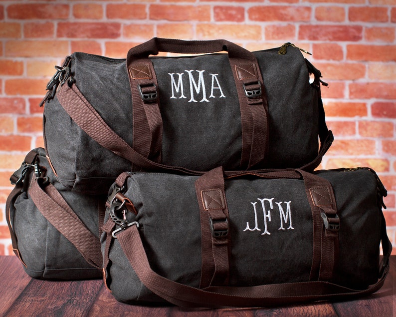 K&ouml;nnte beinhalten: Drei graue Canvas-Reisetaschen mit braunen Lederakzenten und Riemen. Die Taschen sind mit den Initialen "MMA", "JFM" und "JFM" monogrammiert.