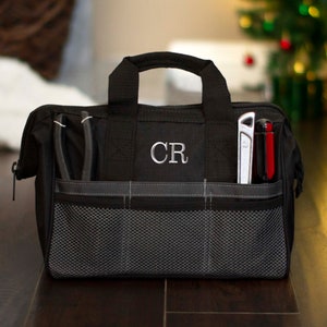 K&ouml;nnte beinhalten: Schwarze Werkzeugtasche mit Rei&szlig;verschluss, einem Netzfach und zwei Seitentaschen. Die Tasche hat einen Griff und die Initialen "CR" sind auf der Vorderseite gestickt.