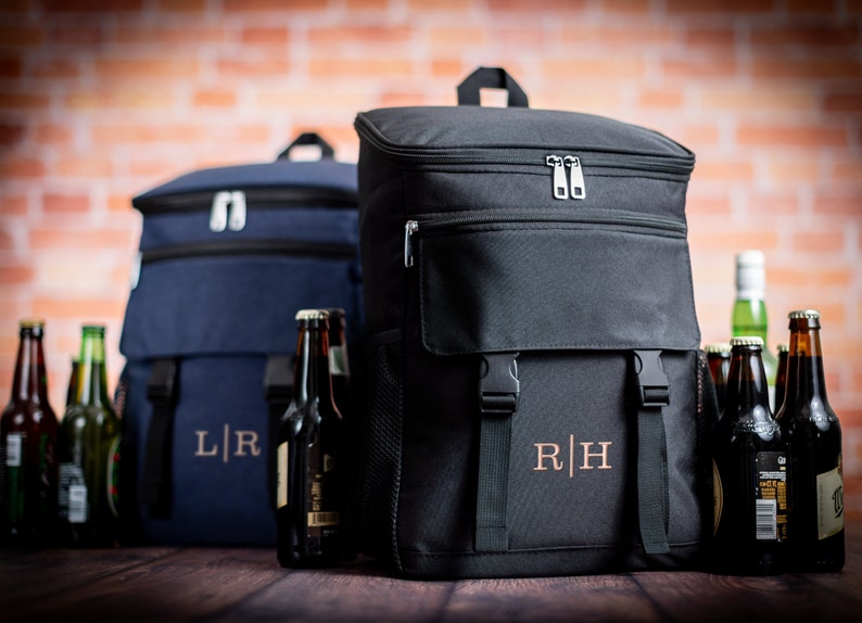 K&ouml;nnte beinhalten: Zwei schwarze und blaue isolierte K&uuml;hlrucks&auml;cke mit verstellbaren Tr&auml;gern und einer Rei&szlig;verschlusstasche vorne. Die Rucks&auml;cke haben personalisierte Initialen, "L|R" und "R|H", die auf der Vorderseite gestickt sind. Die Rucks&auml;cke sind von Bierflaschen umgeben.