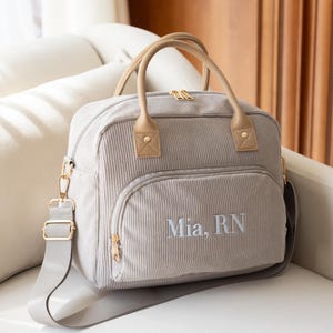 May include: A light gray corduroy diaper bag with a front pocket and top handles. The bag has a shoulder strap and gold-colored hardware. The name "Mia, RN" is embroidered on the front.