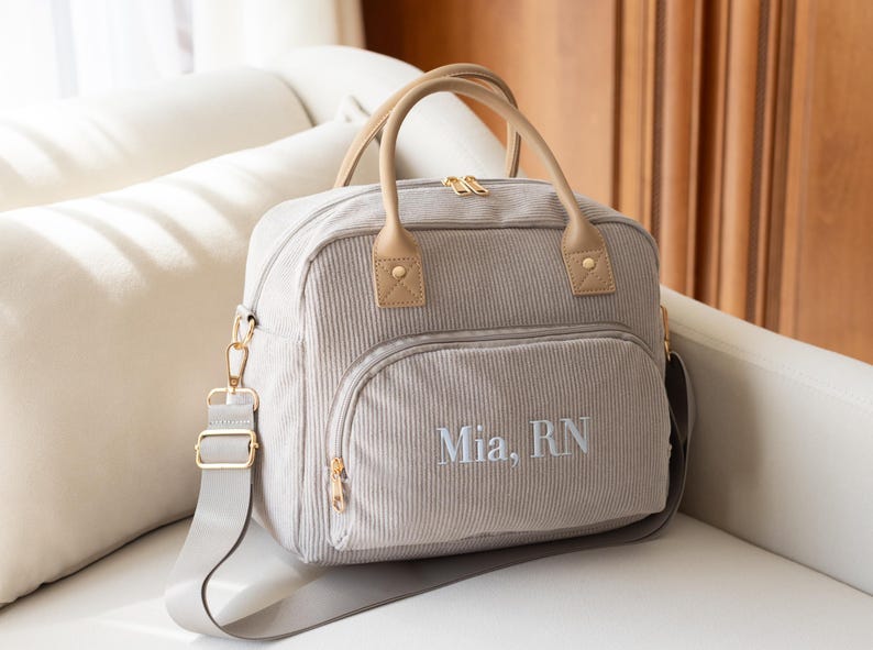 May include: A light gray corduroy diaper bag with a front pocket and top handles. The bag has a shoulder strap and gold-colored hardware. The name "Mia, RN" is embroidered on the front.