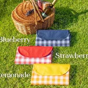Puede incluir: Una cesta de picnic con una botella de vino y comida sobre hierba verde. Se muestran tres mantas de picnic plegadas: Blueberry (a cuadros azules y blancos), Strawberry (a cuadros rojos y blancos) y Lemonade (a cuadros amarillos y blancos).