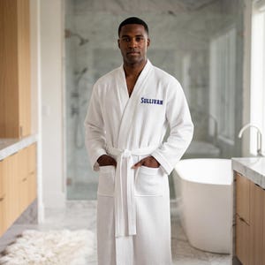 May include: A white waffle-knit bathrobe featuring long sleeves, a tie-waist belt, and two front pockets. The name SULLIVAN is embroidered in blue thread on the left chest area of the robe, which is made of a textured fabric.