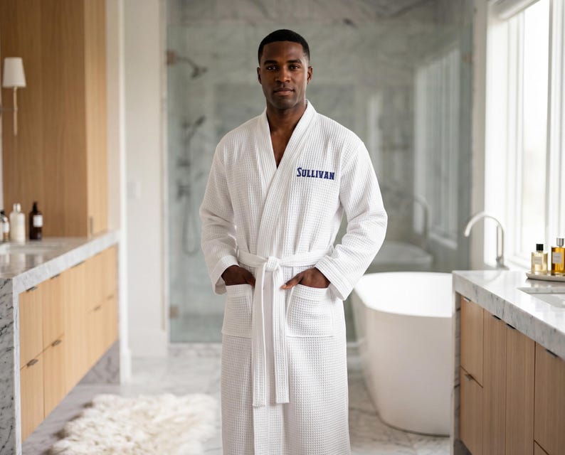 May include: A white waffle-knit bathrobe featuring long sleeves, a tie-waist belt, and two front pockets. The name SULLIVAN is embroidered in blue thread on the left chest area of the robe, which is made of a textured fabric.